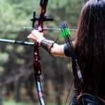 Athletic female aiming with bow and arrow towards the trees